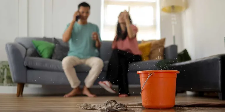 A blurred couple sits on a couch, looking distressed as water leaks from the ceiling into an orange bucket on the floor, with a wet cloth nearby.