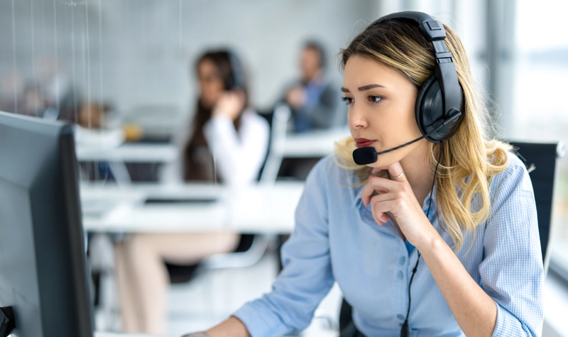 Femme blonde en chemise bleue, portant un casque et regardant un moniteur informatique