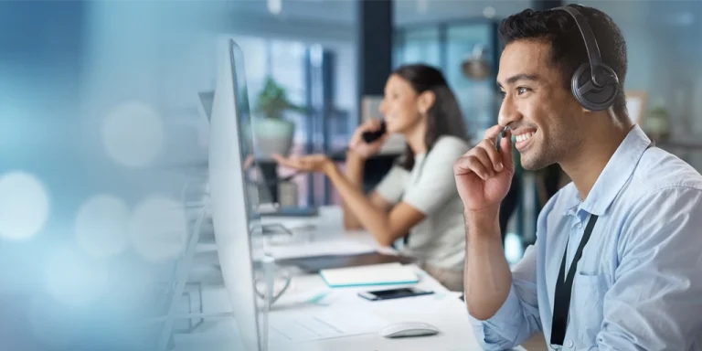 male phone agent wearing a headset, talking to a customer and looking at a computer monitor with female agent in the background