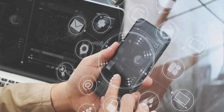 Las manos sosteniendo un teléfono celular rodeadas de iconos tecnológicos con un portátil en el fondo