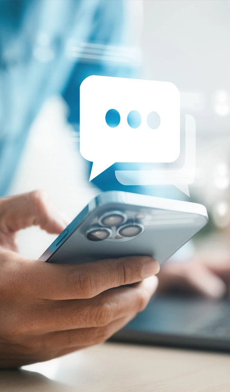 La mano sostiene un teléfono con un icono de mensaje flotante encima y una computadora en segundo plano