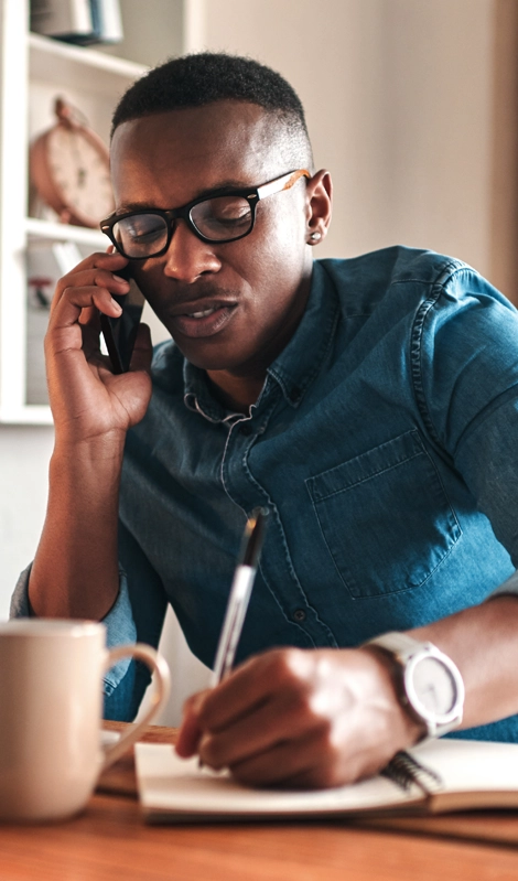 Man i a blue shirt talking on a phone and writing on a notepad